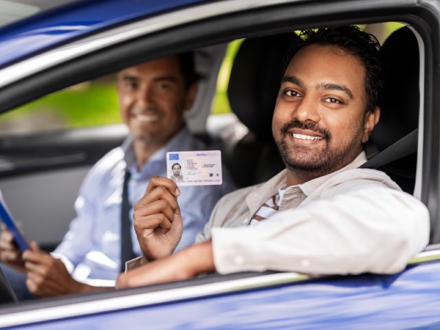 Two men in the car, with the driver holding up his reinstated driver's license with a smile.