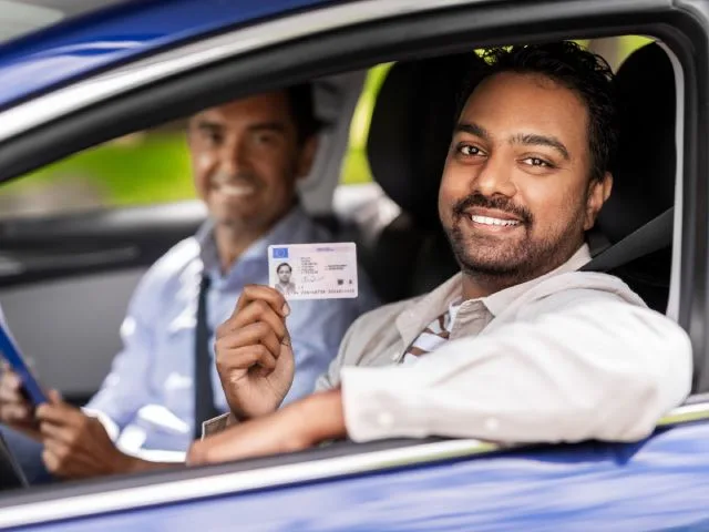 Two men in the car, with the driver holding up his reinstated driver's license with a smile.